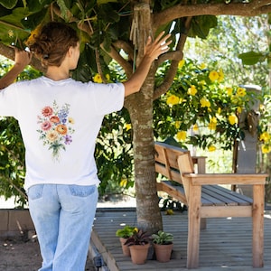 Peut inclure: T-shirt blanc avec un motif de bouquet floral coloré dans le dos. Le t-shirt est associé à un jean bleu clair. La scène se déroule en extérieur avec un banc en bois et de la verdure, dont des fleurs jaunes.