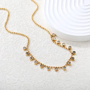 Pode incluir: Um colar dourado com uma corrente delicada e pequenas contas redondas. O colar apresenta v&aacute;rias pedras preciosas redondas e transparentes espa&ccedil;adas ao longo da parte inferior da corrente. Apresentado sobre uma superf&iacute;cie branca.