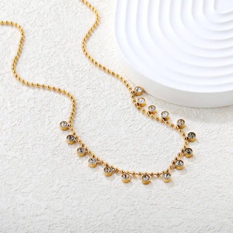 Pode incluir: Um colar dourado com uma corrente delicada e pequenas contas redondas. O colar apresenta v&aacute;rias pedras preciosas redondas e transparentes espa&ccedil;adas ao longo da parte inferior da corrente. Apresentado sobre uma superf&iacute;cie branca.