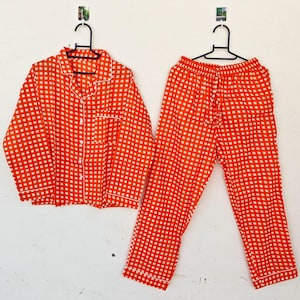 Puede incluir: Conjunto de pijama a cuadros naranja y blanco, que incluye una camisa con botones y pantalones con cordón, colgado contra una pared blanca. La camisa tiene cuello y bolsillo en el pecho. Los bordes del pijama están ribeteados con ribete blanco.