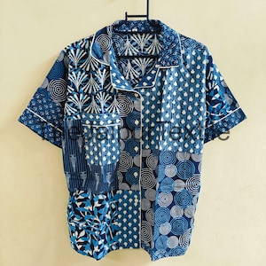 Puede incluir: Una camisa de manga corta con diseño de patchwork en tonos azules y blancos. La camisa presenta varios patrones, incluyendo círculos, puntos y formas de hojas, con ribete blanco a lo largo del cuello y los puños. La camisa está colgada en una percha azul.