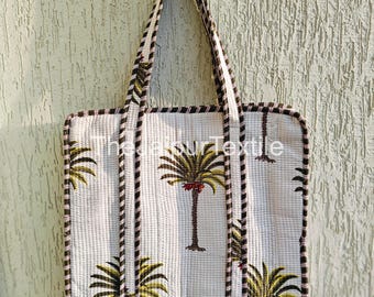 May include: White quilted tote bag with a repeating palm tree pattern in green, brown, and red. The bag has a black and white striped trim and handles. The bag is hanging against a textured, light-colored wall.