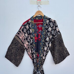 Puede incluir: Una bata kimono estampada con una mezcla de diseños florales negros, rojos y dorados. La bata tiene mangas anchas y un cinturón anudado. La tela parece ligera, adecuada para usar en capas o como abrigo.