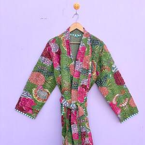 May include: A green patchwork robe with a floral pattern in pink, red, and orange. The robe has long sleeves and a matching belt. The edges are trimmed with a white and green striped fabric. The robe is hanging on a wooden hanger.