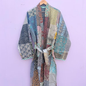 Pode incluir: Robe de patchwork com cinto, pendurado em um cabide de madeira. O robe é feito de vários retalhos de tecido em tons de azul, cinza, laranja e vermelho. As bordas são aparadas com um tecido listrado verde e branco.