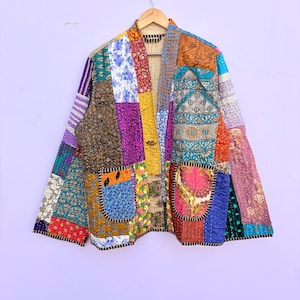 May include: A patchwork jacket with a variety of colorful fabric squares. The jacket has two pockets and is hanging on a wooden hanger. The jacket's design includes floral, geometric, and abstract patterns in shades of blue, purple, orange, and yellow.