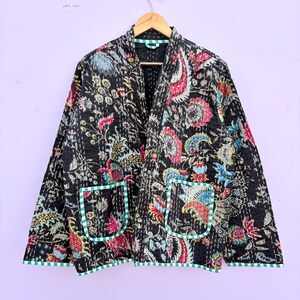 Könnte beinhalten: Eine schwarze Steppjacke mit einem lebendigen Blumenmuster in Rosa, Blau und Grün. Die Jacke hat zwei Vordertaschen mit grün-weißem Streifenbesatz und hängt an einem Holzkleiderbügel.