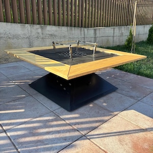 Può includere: Un tavolo braciere quadrato da esterno con una cornice in legno chiaro che circonda un centro in metallo nero. L'unità include una griglia centrale in metallo nero e una griglia di cottura rialzata in metallo argentato, su una base nera.