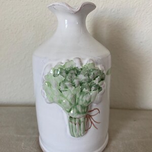 May include: White ceramic vase with a ruffled top and a painted bouquet of green lettuce. The bouquet is tied with a pink ribbon. The vase has a slightly distressed, rustic appearance.