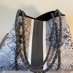 Könnte beinhalten: Eine große Tragetasche mit Schlangenmuster in Grau- und Weißtönen. Die Tasche hat einen zentralen grauen Streifen, der von weißen Streifen flankiert wird, und schwarze Seilgriffe mit weißen Akzenten. Der obere Rand ist schwarz.