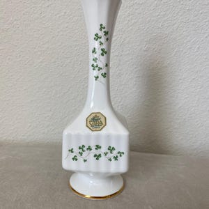 May include: White ceramic vase with a gold rim and base. The vase features green shamrock designs and the text "Royal Tara China". The vase has a unique shape with a flared top and a square base.