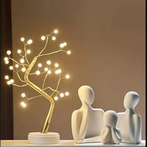 Puede incluir: Una lámpara decorativa dorada en forma de árbol con pequeñas luces blancas cálidas. La lámpara se apoya sobre una base blanca. Tres figuras humanas abstractas blancas están a la derecha de la lámpara.