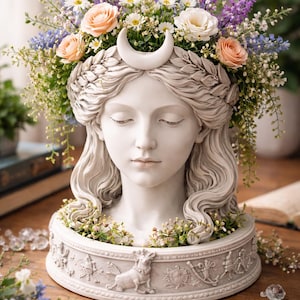 May include: A white sculpted head of a woman with closed eyes, decorated with a crescent moon and a floral crown. The flowers include peach roses, white daisies, and purple and blue wildflowers. The base has a detailed relief design.