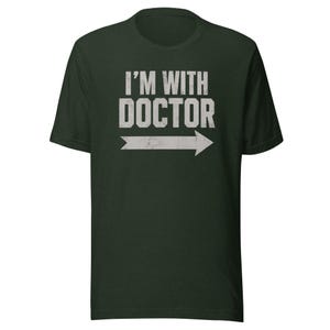 May include: A dark forest green short-sleeve t-shirt featuring the white distressed text "I'M WITH DOCTOR" printed on the chest. Below the text is a white horizontal arrow pointing to the right, also displaying a distressed, weathered texture.