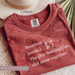May include: A red Comfort Colors t-shirt with the text "Bourbon (noun.) The glue holding this shit show together" and a graphic of a bourbon glass. The shirt also has the text "More colours & Styles".