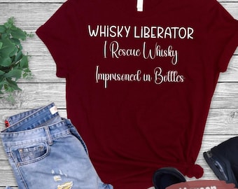 May include: A maroon t-shirt featuring the white text "WHISKY LIBERATOR I Rescue Whisky Imprisoned in Bottles". The shirt is displayed with blue jeans and black shoes. The shirt is a solid colour.