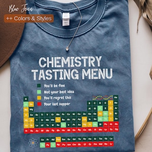 Op de afbeelding: Blauw T-shirt met de witte tekst "CHEMISTRY TASTING MENU". Het ontwerp bevat een periodiek systeem met kleurgecodeerde elementen en zinnen als "You'll be fine" en "Your last supper".