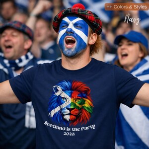 May include: Navy blue t-shirt with a colorful lion graphic split into halves, one side with the Scottish flag design and the other in rainbow colors. The text "Scotland is the Party 2026" is below the graphic. A person is wearing the shirt and has their face painted with the Scottish flag.