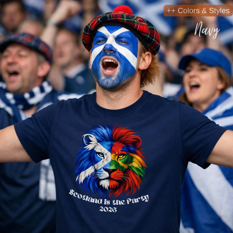 May include: Navy blue t-shirt with a colorful lion graphic split into halves, one side with the Scottish flag design and the other in rainbow colors. The text "Scotland is the Party 2026" is below the graphic. A person is wearing the shirt and has their face painted with the Scottish flag.