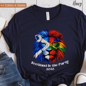 May include: Navy blue t-shirt featuring a colorful lion design. The lion's face is split, with one side displaying the Scottish flag and the other side a vibrant rainbow of colors. The text "Scotland is the Party 2026" is printed below the lion.