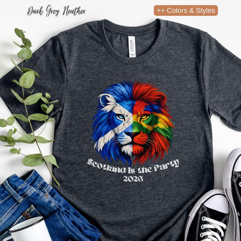 May include: Dark grey heather t-shirt featuring a colorful lion graphic. The lion's face is split, with one side displaying the Scottish flag and the other side a rainbow of colors. Text on the shirt reads "Scotland is the Party 2026."