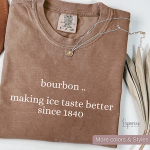 May include: A brown Comfort Colors t-shirt with the text "bourbon .. making ice taste better since 1840" in white. A silver necklace with a small pendant is draped over the shirt. The shirt is size small.
