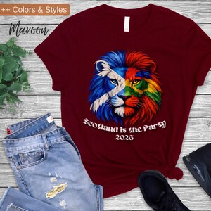 May include: Maroon t-shirt featuring a lion design split with the Scottish flag and rainbow colors. The text "Scotland is the Party 2026" is printed below the lion. The shirt is paired with blue jeans and black sneakers.