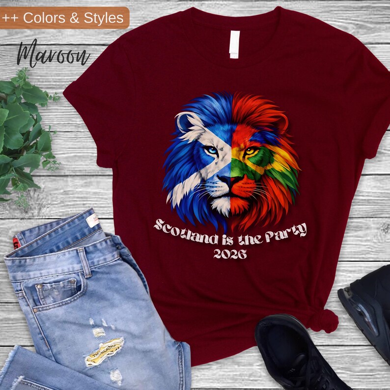 May include: Maroon t-shirt featuring a lion design split with the Scottish flag and rainbow colors. The text "Scotland is the Party 2026" is printed below the lion. The shirt is paired with blue jeans and black sneakers.