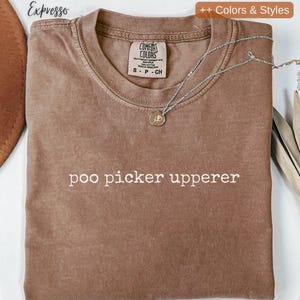 May include: A brown Comfort Colors t-shirt with the phrase "poo picker upperer" printed in white. A silver necklace with a circular pendant rests on the shirt. The shirt is folded and has a tag at the neckline.