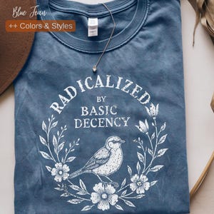 Puede incluir: Una camiseta azul con el texto "RADICALIZED BY BASIC DECENCY" en un diseño de corona floral blanca. Una ilustración detallada de un pájaro está centrada debajo del texto. La camiseta tiene cuello redondo y mangas cortas.