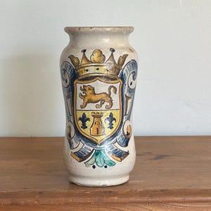 May include: A beige ceramic vase with a heraldic crest design. The crest features a golden lion, fleur-de-lis, and a castle, set against a blue and yellow shield. The vase is cylindrical with a slightly flared top, and stands on a wooden surface.
