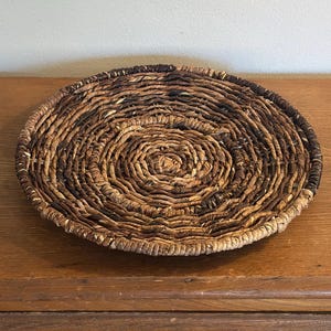 May include: A round, woven basket with a spiral design. The basket is made of natural-colored fibers, with darker brown accents. The basket is sitting on a wooden surface.