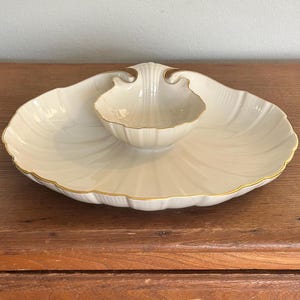 May include: A cream-colored ceramic serving dish shaped like a large scallop shell with a smaller, integrated shell-shaped bowl in the center. The edges of both the large dish and the central bowl are trimmed with a thin, metallic gold-colored line.
