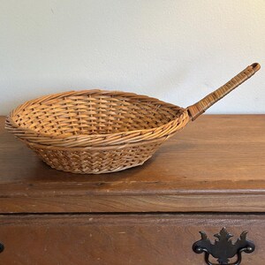 May include: A woven wicker basket with a long handle, resembling a small frying pan. The basket is light brown and sits on a wooden surface. The handle is also woven and extends from the side of the basket.
