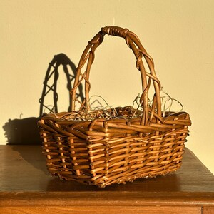May include: A rectangular woven wicker basket with a handle. The basket is made of light brown wicker and has a sturdy handle. The basket is sitting on a wooden surface. The basket is empty.