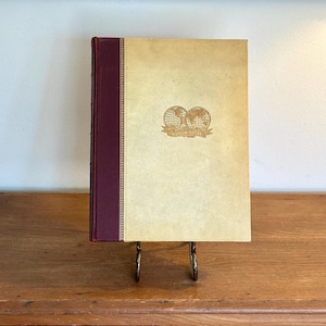 May include: A vintage book with a tan cover and a burgundy spine, displayed on a decorative stand. The cover features a gold emblem of two globes and the words "World Atlas". The book is set against a wooden dresser.