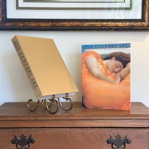 May include: A still life featuring two books on a wooden surface. One book, titled "19th-CENTURY ART," is propped up on a decorative metal stand. The other book, also titled "19th-CENTURY ART," displays a painting of a woman in an orange dress.