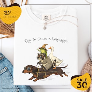 a white t-shirt with a whimsical illustration of a dog, frog, and duck riding on a skateboard, accompanied by the text "Off to Cause a Kerfluffle". The t-shirt is displayed on a white background, and there are yellow and black sale signs indicating a 30% discount.