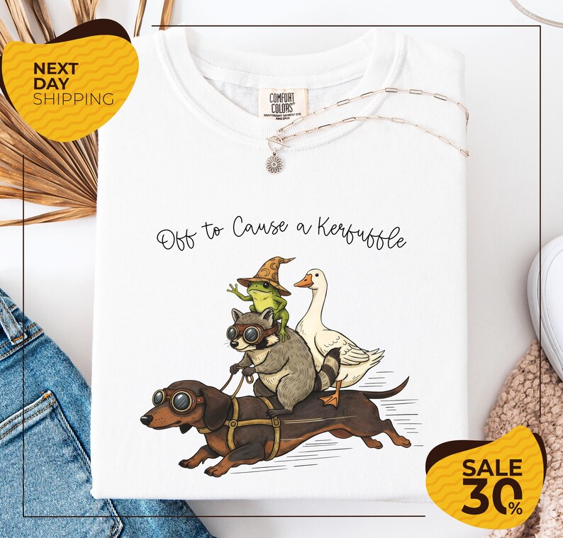 a white t-shirt with a whimsical illustration of a dog, frog, and duck riding on a skateboard, accompanied by the text "Off to Cause a Kerfluffle". The t-shirt is displayed on a white background, and there are yellow and black sale signs indicating a 30% discount.