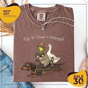 a brown t-shirt with a graphic design depicting a dog, frog, and duck riding on a bicycle, along with the text "Off to Cause a Kerfluffle". The t-shirt is displayed on a white background, and there are yellow and black sale signs indicating a 30% discount.