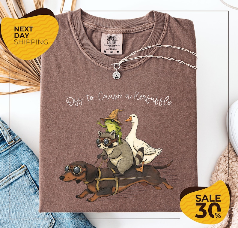 a brown t-shirt with a graphic design depicting a dog, frog, and duck riding on a bicycle, along with the text "Off to Cause a Kerfluffle". The t-shirt is displayed on a white background, and there are yellow and black sale signs indicating a 30% discount.