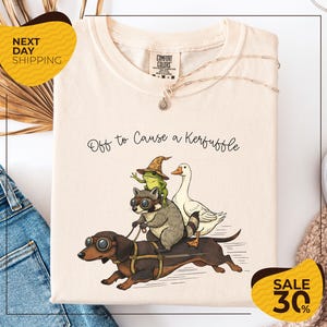 a white t-shirt with a whimsical illustration of a frog, a dog, and a duck riding on a dachshund, accompanied by the text "Off to Cause a Kerfluffle". The t-shirt is displayed on a white background, and there are two yellow sale signs indicating a 30% discount on the item.