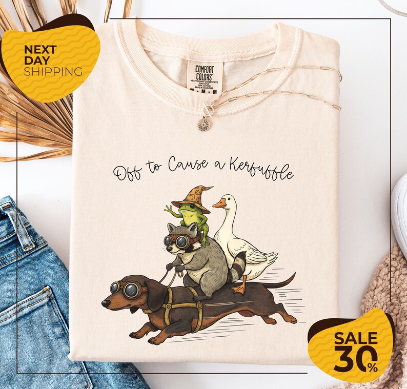 a white t-shirt with a whimsical illustration of a frog, a dog, and a duck riding on a dachshund, accompanied by the text "Off to Cause a Kerfluffle". The t-shirt is displayed on a white background, and there are two yellow sale signs indicating a 30% discount on the item.