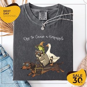 a gray t-shirt with a whimsical graphic design depicting a dog, a frog, a duck, and a raccoon riding on a dachshund. The text on the t-shirt reads "Off to Cause a Kerfluffle". The t-shirt is displayed on a white background, and there are two yellow sale signs in the image, one on the left and one on the right, indicating a 30% discount.