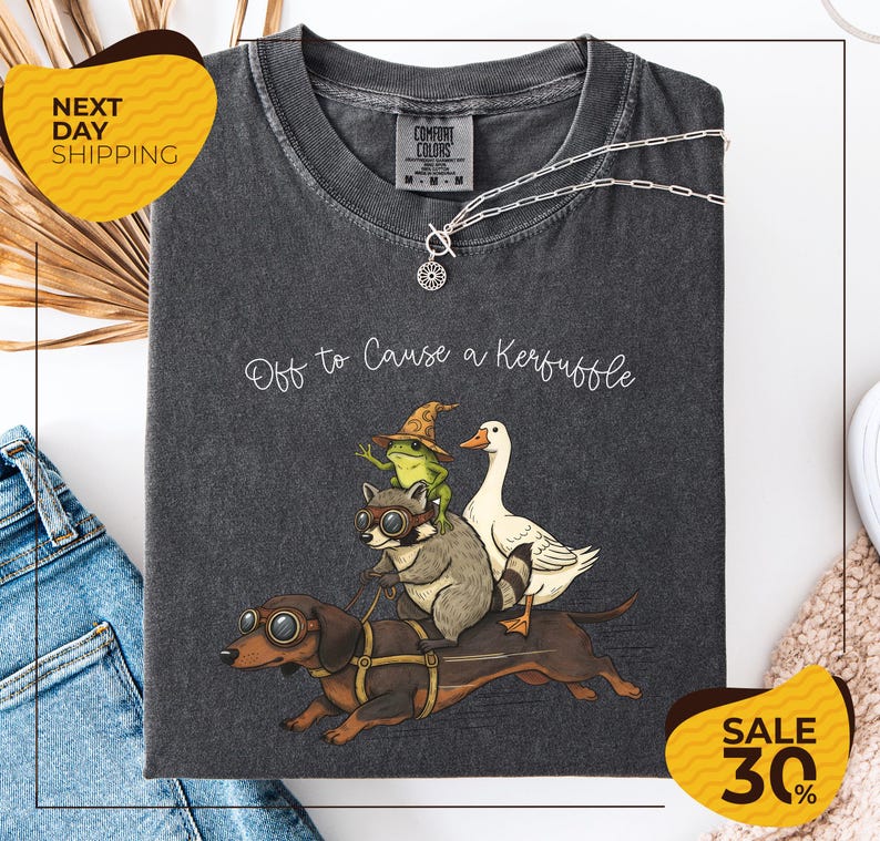 a gray t-shirt with a whimsical graphic design depicting a dog, a frog, a duck, and a raccoon riding on a dachshund. The text on the t-shirt reads "Off to Cause a Kerfluffle". The t-shirt is displayed on a white background, and there are two yellow sale signs in the image, one on the left and one on the right, indicating a 30% discount.