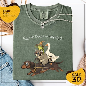 a green t-shirt with a cartoon illustration of a dog, cat, frog, and duck riding on a dachshund, accompanied by the text "Off to Cause a Kerfluffle". The t-shirt is displayed on a white background, and there are yellow and black sale signs indicating a 30% discount.