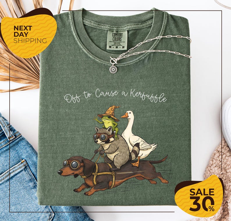 a green t-shirt with a cartoon illustration of a dog, cat, frog, and duck riding on a dachshund, accompanied by the text "Off to Cause a Kerfluffle". The t-shirt is displayed on a white background, and there are yellow and black sale signs indicating a 30% discount.