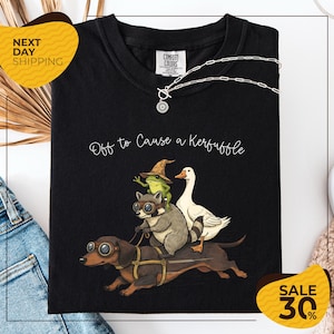 a black t-shirt with a whimsical graphic design depicting a dog, a frog, a duck, and a raccoon riding on a dog's back. The text on the t-shirt reads "Off to Cause a Kerfluffle". The t-shirt is displayed on a white background, and there are yellow and black sale signs indicating a 30% discount.