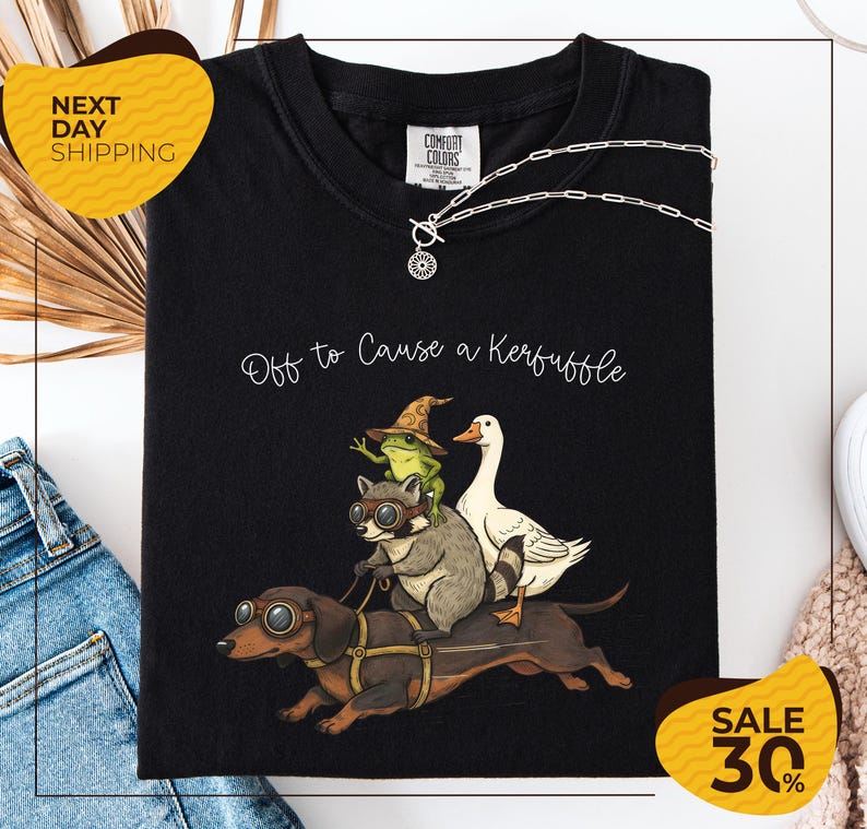 a black t-shirt with a whimsical graphic design depicting a dog, a frog, a duck, and a raccoon riding on a dog's back. The text on the t-shirt reads "Off to Cause a Kerfluffle". The t-shirt is displayed on a white background, and there are yellow and black sale signs indicating a 30% discount.