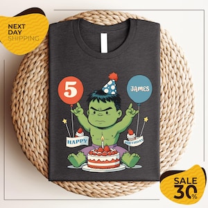 A t-shirt with a graphic design depicting a cartoon character wearing a party hat and holding a birthday cake, with balloons and the text "Happy Birthday James" around the character.